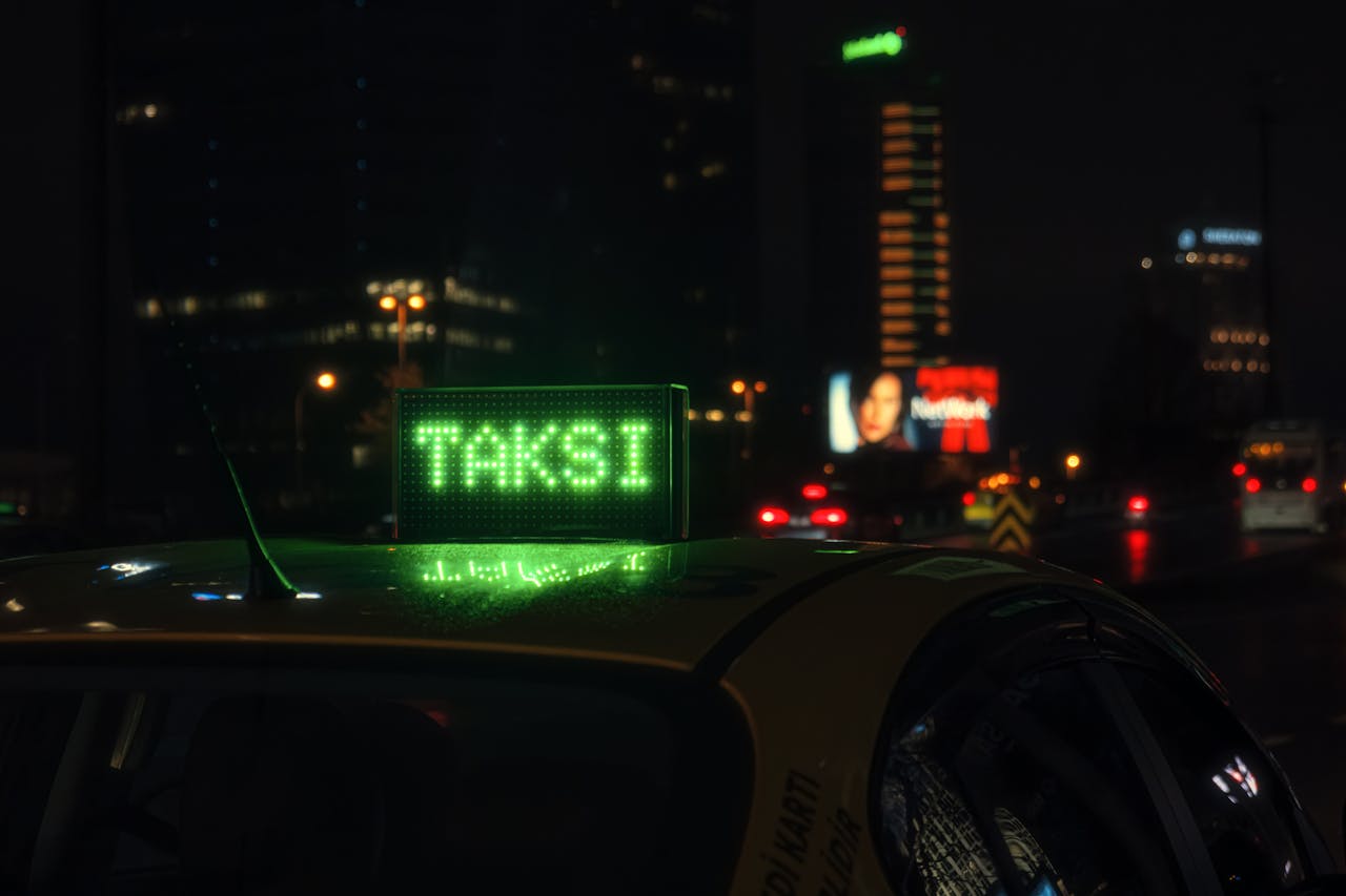 Nighttime cityscape with a glowing 'TAKSI' sign on a taxi roof, vibrant urban setting.